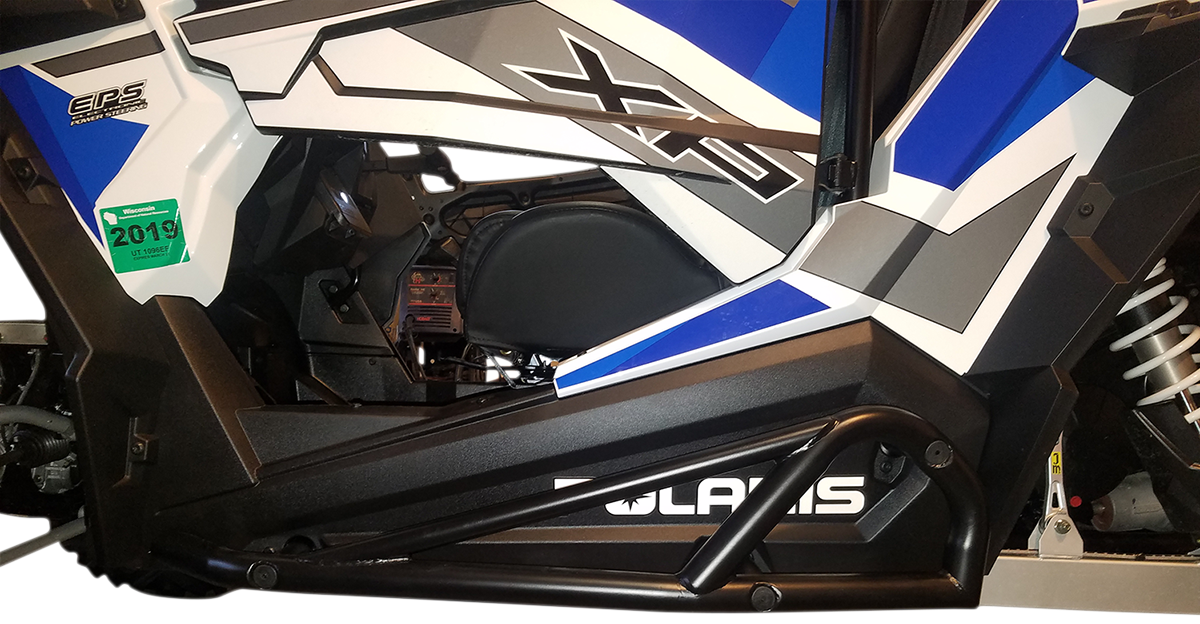 A close-up side view of a Polaris off-road vehicle features angular black, white, gray, and blue body panels, a green 2019 registration sticker, and visible suspension components. Polaris is partially visible on the lower panel.
