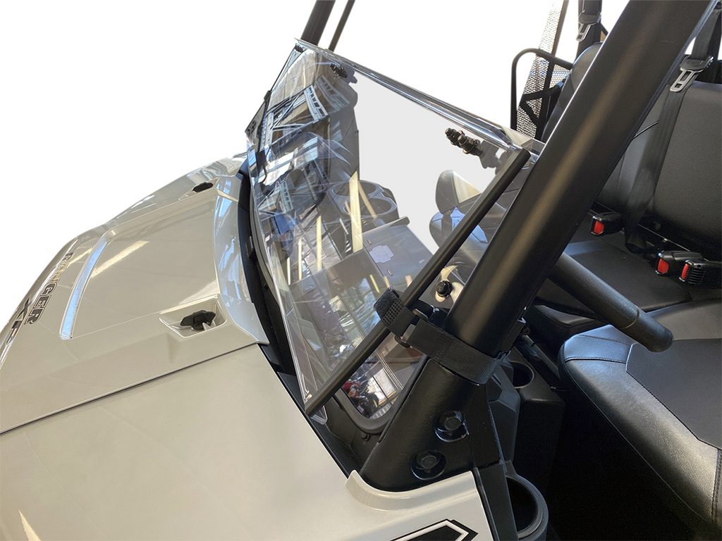 A close-up view of a clear, slightly angled windshield mounted on a white off-road utility vehicle. The windshield is attached to sturdy black bars, and the interior shows two black seats with seat belts. The background is plain and white.