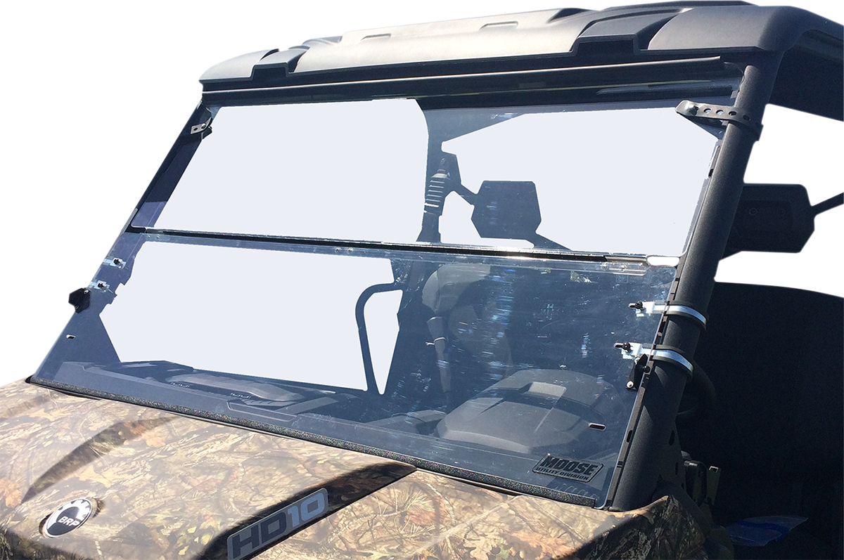 A clear, two-piece polycarbonate windshield is mounted on a camouflage-patterned utility vehicle. The windshield is held in place by metal hinges and brackets, and the background is completely white, highlighting the windshield and vehicle details.