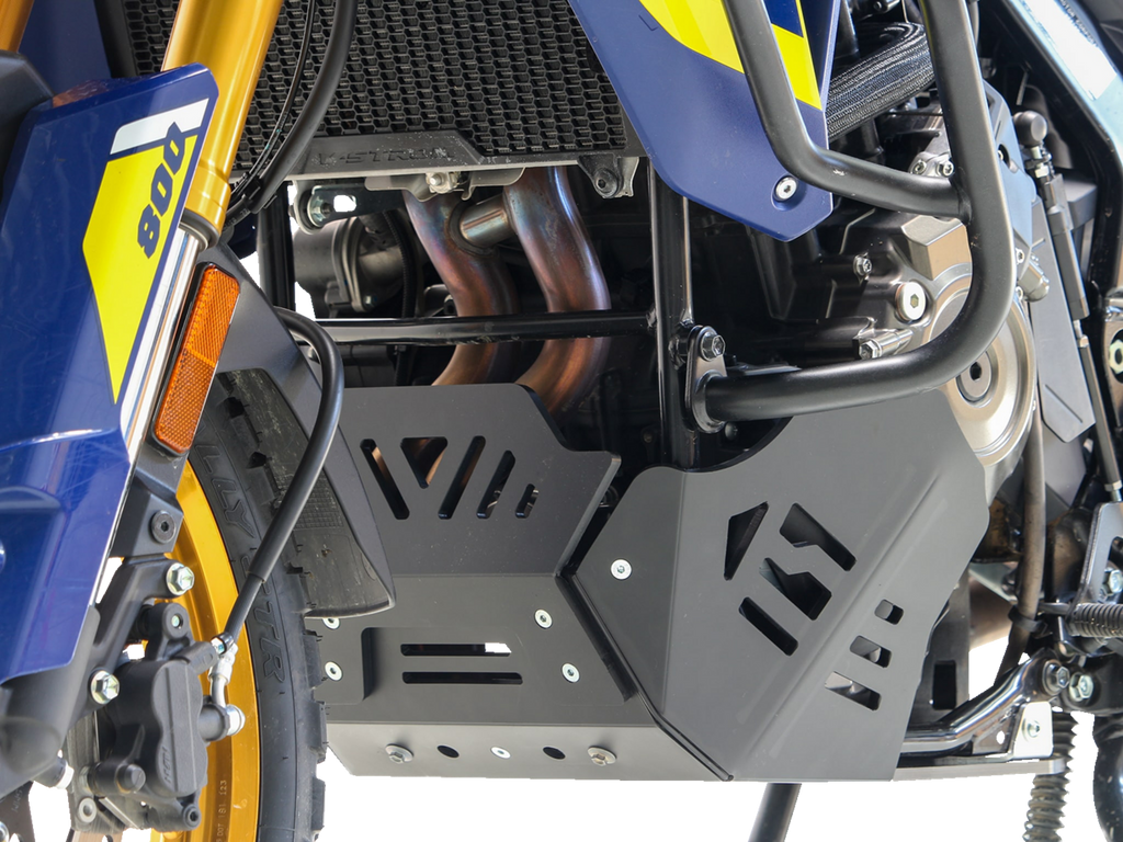 Close-up of a blue and yellow motorcycle’s lower section, highlighting the black skid plate protecting the engine and exhaust pipes. The front suspension fork, part of a wheel, and some engine components are also visible, with intricate metal and plastic 