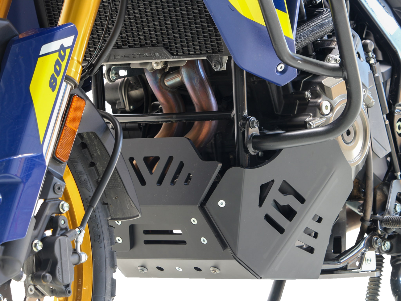 Close-up of a blue and yellow motorcycle’s lower section, highlighting the black skid plate protecting the engine and exhaust pipes. The front suspension fork, part of a wheel, and some engine components are also visible, with intricate metal and plastic 