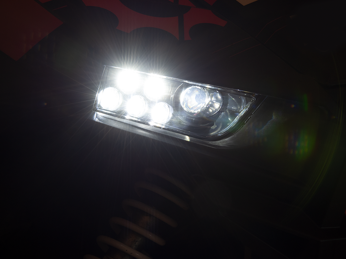 A close-up of a bright car headlight with multiple LED bulbs shining white light. The light beams create lens flares, and the suspension coil is faintly visible below the headlight in the shadowy background.