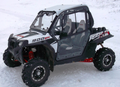 A Polaris RZR XP900/900s Cab Kit is installed on a black and white off-road side-by-side, shown on snow with rugged tires, red suspension springs, two bucket seats, and clear cab doors providing partial enclosure.