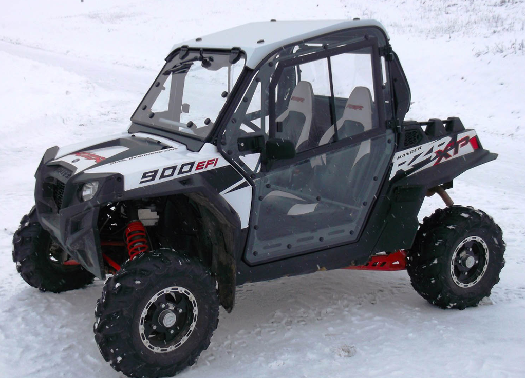 A Polaris RZR XP900/900s Cab Kit is installed on a black and white off-road side-by-side, shown on snow with rugged tires, red suspension springs, two bucket seats, and clear cab doors providing partial enclosure.