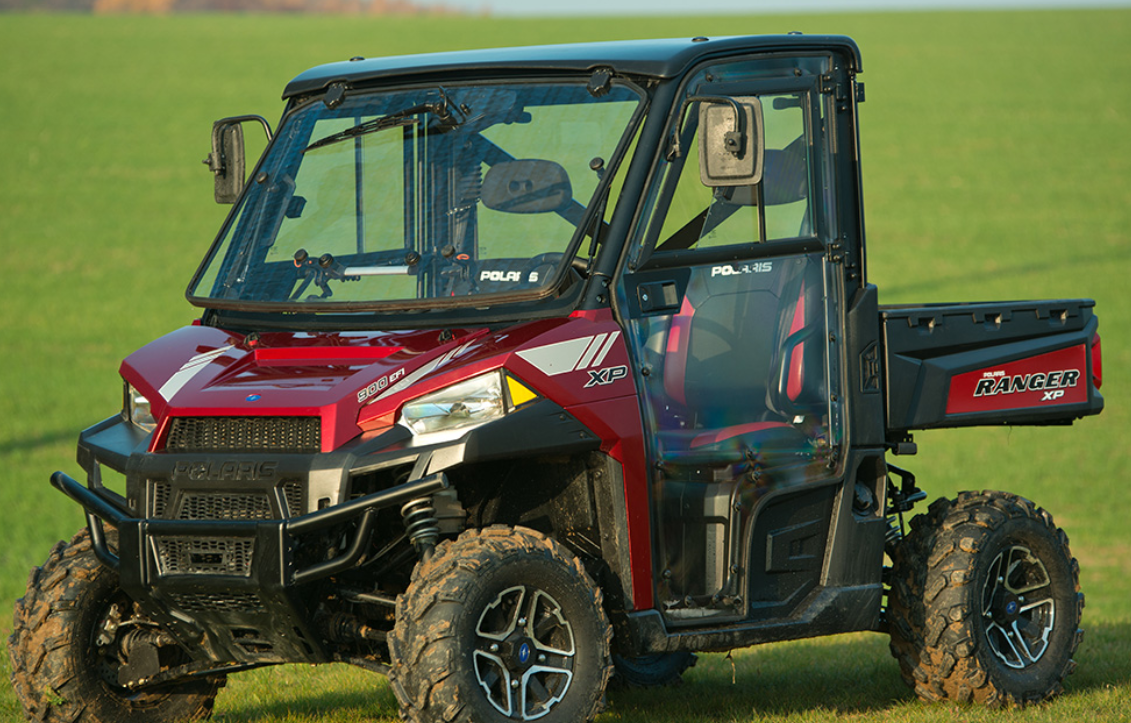 A Polaris Ranger XP900 (2013–PRESENT) with a Full Hard Cab Enclosure, clear doors, and large off-road tires is parked on green grass. The side cargo bed features Ranger XP branding; a blurred field and sky are in the background.