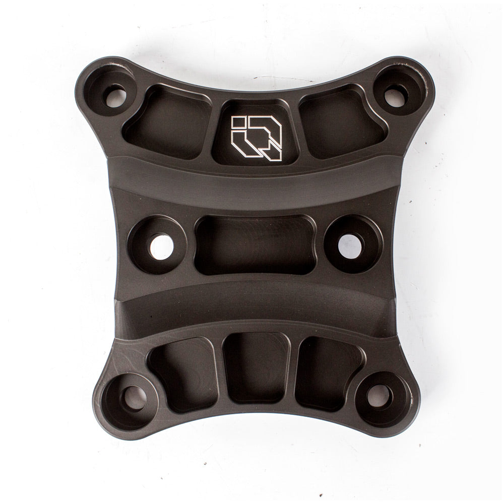 The Can Am Maverick X3 / Max HMF Billet Back Plate Black (2017+) is a black, machined metal plate with four rounded corners, five countersunk holes, and a central logo of stylized geometric shapes, shown on a white background.