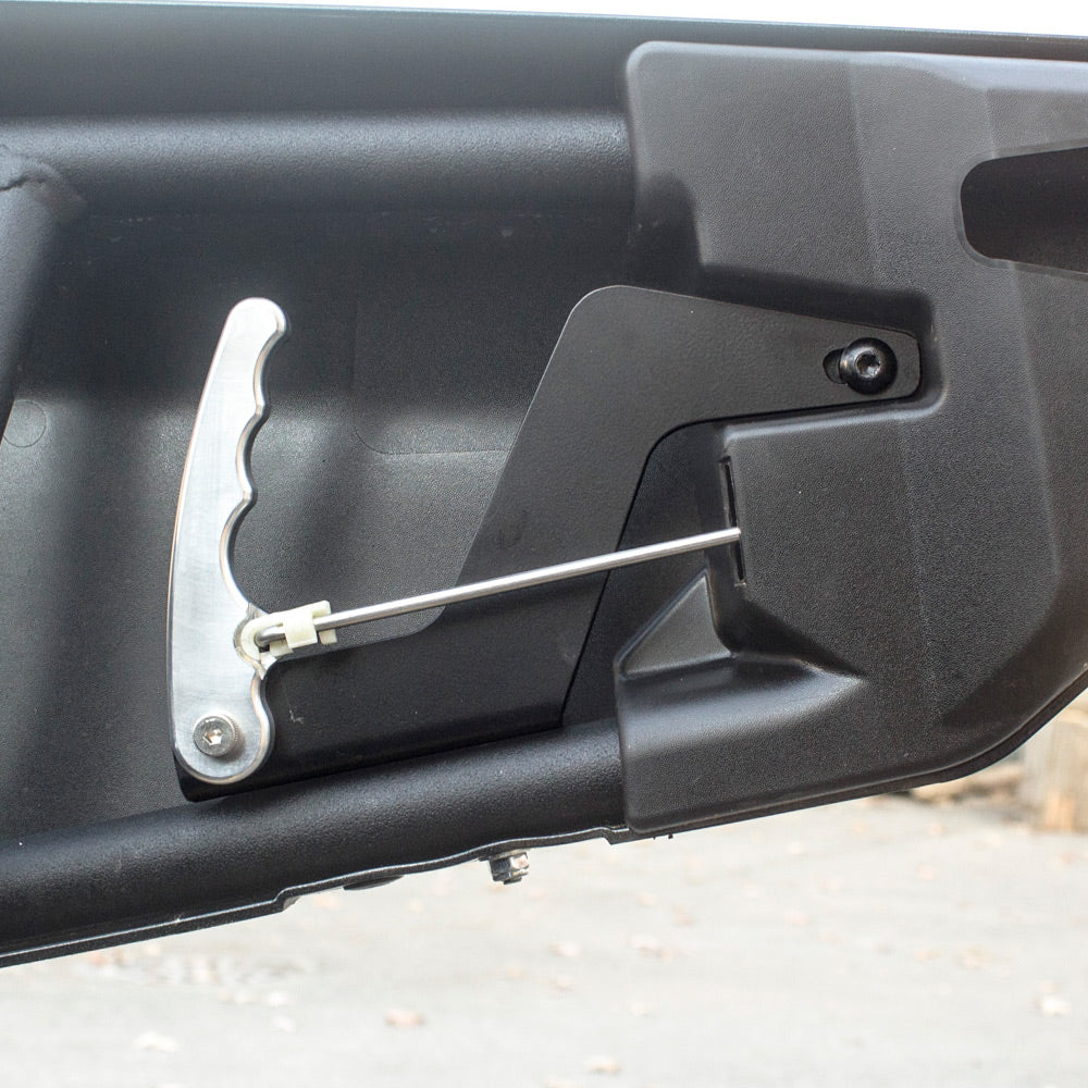 A close-up of the Can Am Maverick X3 HMF Easy-Grip Aluminum Door Handle (2017+) and rod mechanism installed inside a black vehicle tailgate, highlighting the handle, rod, and mounting hardware against the plastic inner panel.