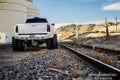 A white lifted pickup with off-road tires, fitted with a 2010-2018 Ram 2500 Stealth Rear Winch Bumper (Texture Black, Warn M8000 only), is parked on gravel by train tracks with hills and industrial tanks in the background; watermark at lower right.