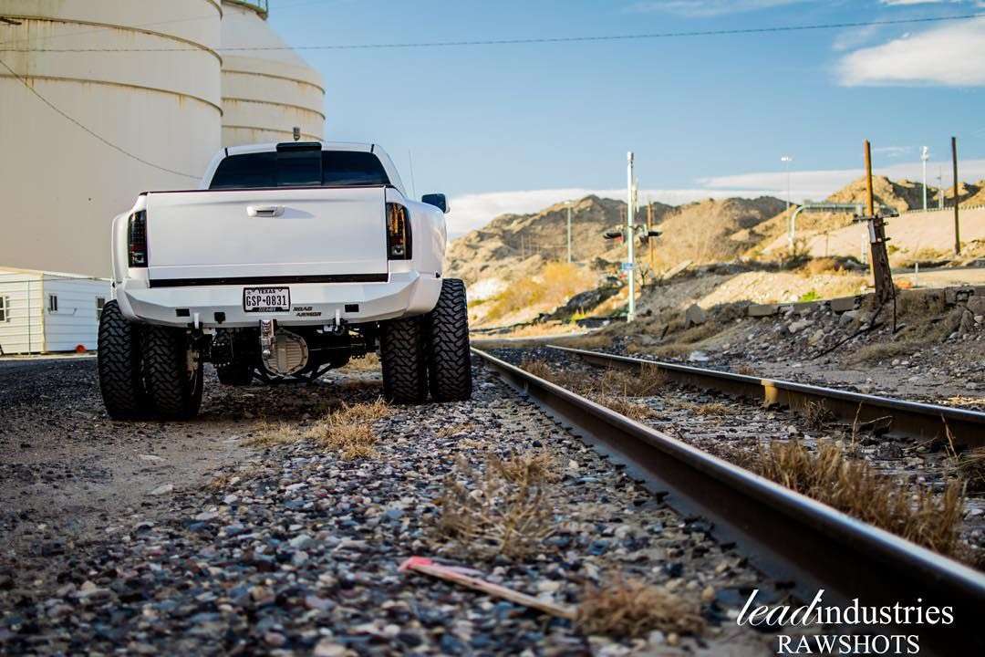 A white lifted pickup with off-road tires, fitted with a 2010-2018 Ram 2500 Stealth Rear Winch Bumper (Texture Black, Warn M8000 only), is parked on gravel by train tracks with hills and industrial tanks in the background; watermark at lower right.