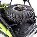 A Honda Talon 1000R/X HMF Tire Rack (Black, 2019+) is mounted on the rear of a green utility vehicle, securely holding a large off-road spare tire with deep treads, visible black straps, and a center ratchet strap for added security.