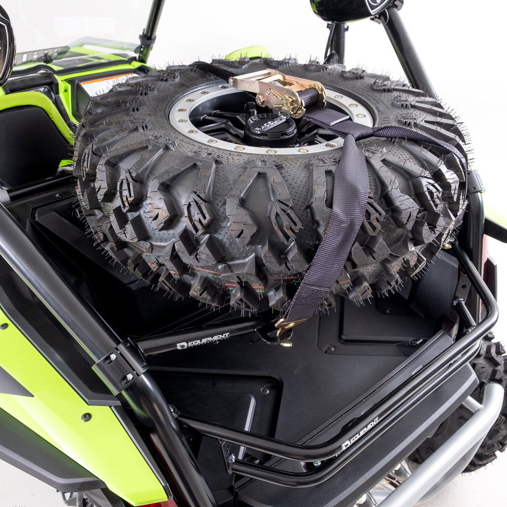A Honda Talon 1000R/X HMF Tire Rack (Black, 2019+) is mounted on the rear of a green utility vehicle, securely holding a large off-road spare tire with deep treads, visible black straps, and a center ratchet strap for added security.
