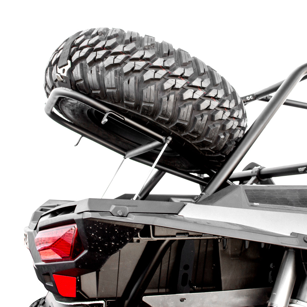 A close-up of the Polaris Rzr Turbo | Rzr Xp1K Hmf Spare Tire Rack - Black 2014-23 shows a rugged spare tire mounted on an angled rack above the cargo bed, with red taillight, roll cage, and frame visible against a white background.