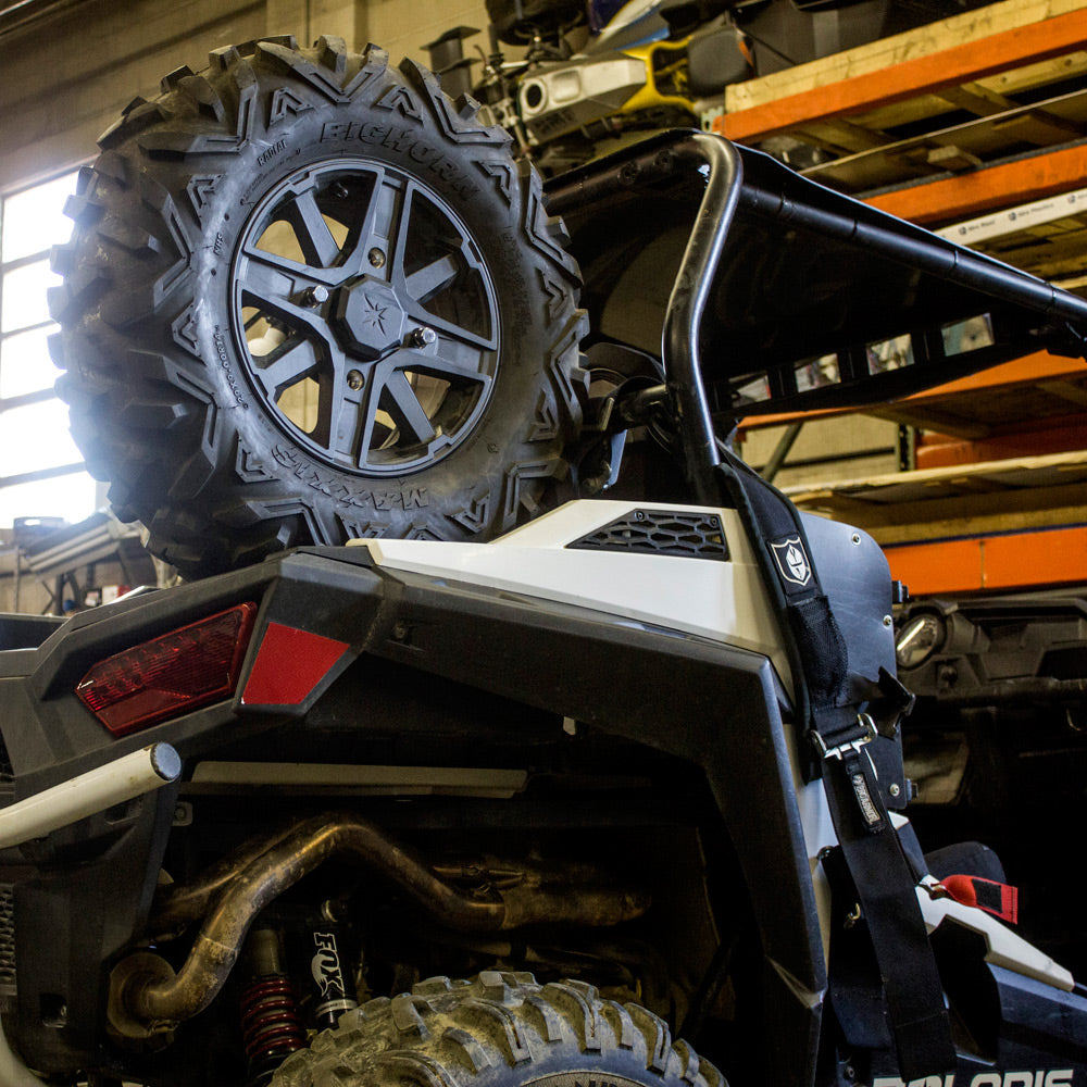A close-up shows the Polaris Rzr S1000 / S900 HMF Spare Tire Rack - Black (2015+) mounted on the rear of an off-road vehicle, with a spare tire and garage shelves with tools visible in the background.