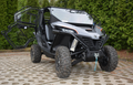A rugged CF MOTO Zforce 950 Full Cab Enclosure (2020-Present) with large knobby tires and open side doors is parked on patterned concrete, featuring a front bumper winch and surrounded by tall green trees.