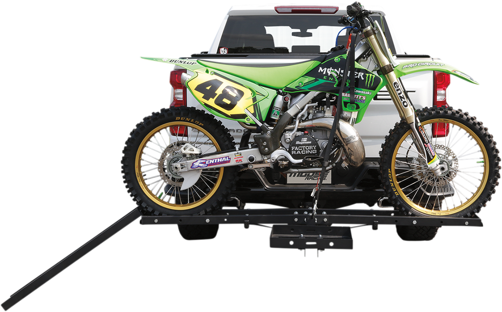 A green and yellow motocross dirt bike with the number 48 is mounted on a black hitch rack attached to the rear of a white pickup truck. The bike is secured, and a ramp extends from the rack to the ground for loading and unloading.
