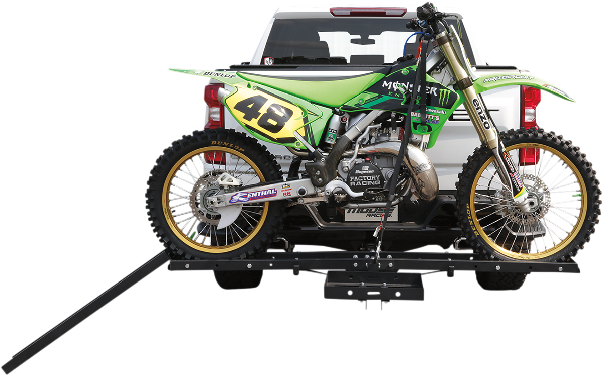 A green and yellow motocross dirt bike with the number 48 is mounted on a black hitch rack attached to the rear of a white pickup truck. The bike is secured, and a ramp extends from the rack to the ground for loading and unloading.