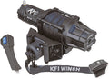 A black 5000 Assault Series Winch with wrapped cable, mounted bracket, and attached hook. At left is a handheld remote with blue buttons; at right are a “KFI WINCH” strap and a wired control switch.