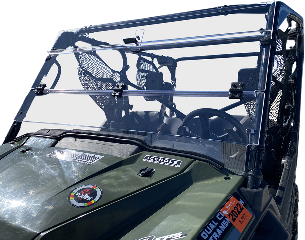A close-up of a green off-road utility vehicle shows a clear two-piece windshield with black mounting brackets. The dashboard features various stickers, including ICEHOLE and Duel Club Frame 2022. The seats and a mesh storage pouch are visible inside.