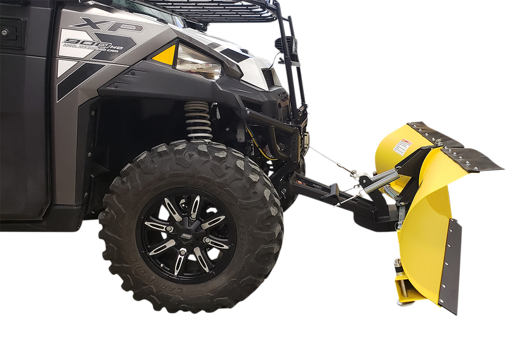 A close-up side view of an off-road utility vehicle with rugged black tires, featuring a large, curved yellow snow plow attachment mounted to its front. The plow is angled and has a black edge for scraping, with metal supports connecting it to the vehicle