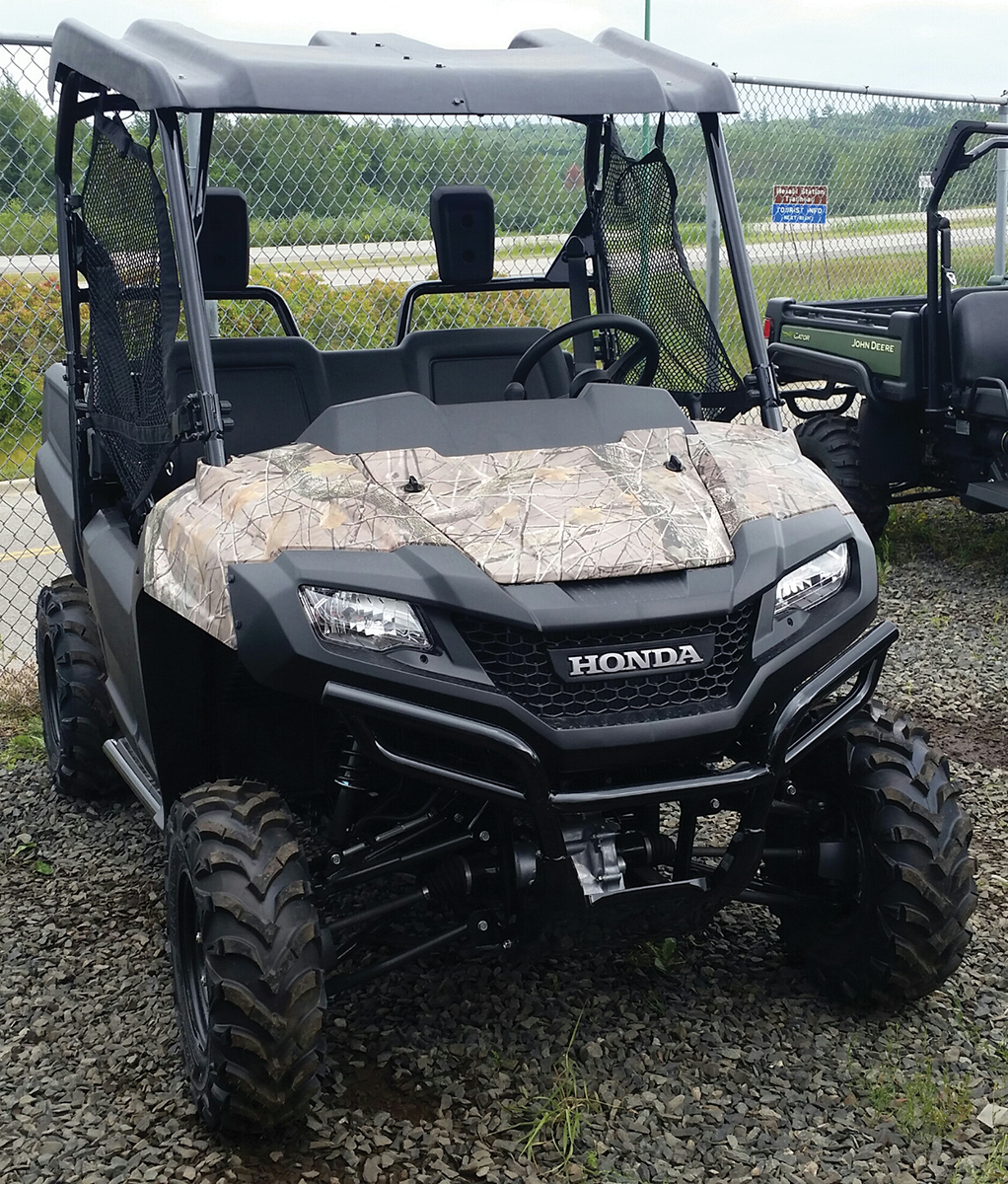 A camouflage-patterned Honda utility vehicle with roll cage, rugged off-road tires, and open sides is parked on gravel near a chain-link fence. Another green utility vehicle is partially visible in the background. Trees and a road are in the distance.