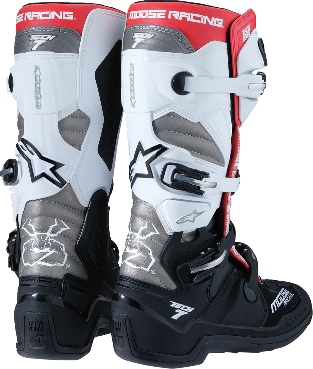 A pair of Moose Racing motocross boots, primarily black, white, and silver, with red accents. The boots feature multiple buckles, textured panels, and bold logos, offering ankle and shin protection for off-road motorcycle riding.