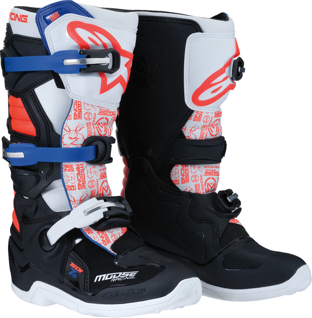 A pair of motocross boots with black, white, red, and blue accents. They feature bold graphics, metal buckles, protective panels, and reinforced soles. The boots have prominent branding and are designed for off-road motorcycle riding.