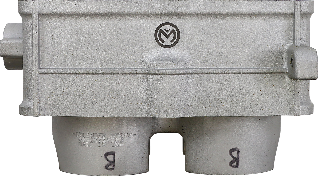 Close-up of a metallic industrial part with a light gray finish. The part has two cylindrical outlets at the bottom, each marked with the letter B. A round logo with a stylized M is centered on the raised section above the outlets.