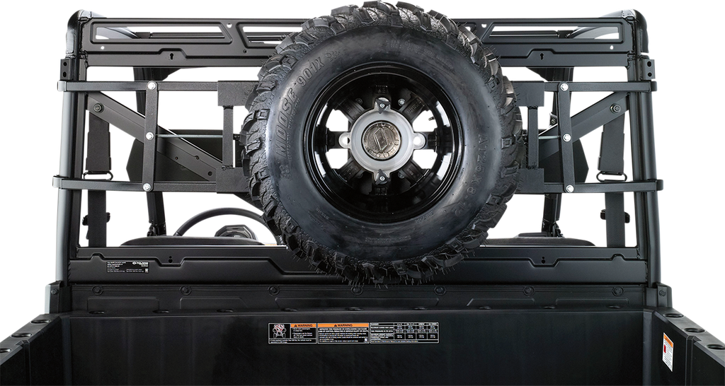 A black spare tire mounted vertically on a metal rack at the back of a utility vehicle bed, with visible bolts and support bars. Labels and safety instructions are attached below the tire on the black bed wall. The background is plain white.
