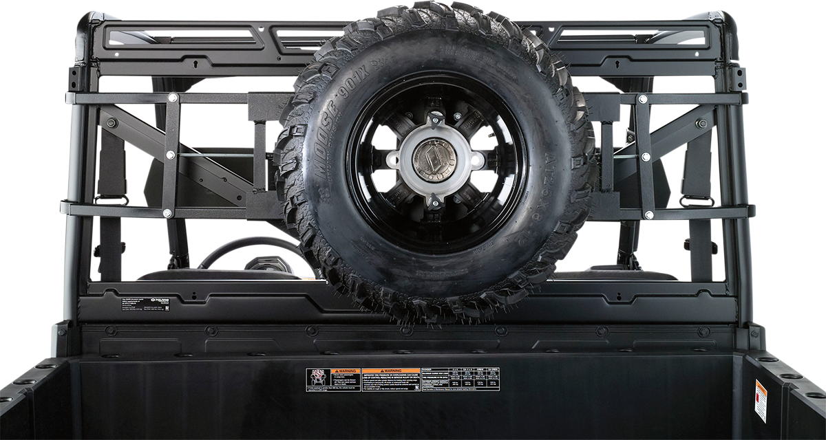 A black spare tire mounted vertically on a metal rack at the back of a utility vehicle bed, with visible bolts and support bars. Labels and safety instructions are attached below the tire on the black bed wall. The background is plain white.