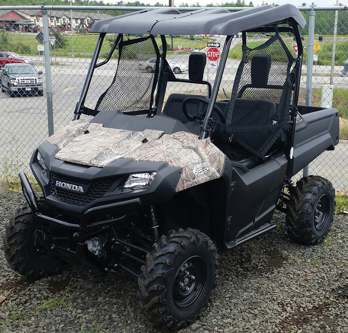 A camouflaged Honda utility terrain vehicle (UTV) with off-road tires is parked on gravel near a chain-link fence. The vehicle has two seats, a mesh windshield, and a cargo bed. A road, cars, and a stop sign are visible in the background.