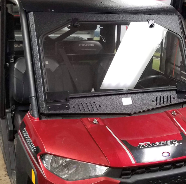 A Polaris Ranger utility vehicle with a black Polaris Ranger 1000 Front Glass Windshield (2019+) is shown in a garage. The windshield features mounted brackets, and the 1000 and RANGER labels are visible on the vehicles hood and front.