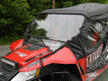 A close-up of a red and black Wildcat X featuring an Arctic Cat Wildcat Split Lexan Windshield (2012-2018) and partial soft top, parked on a paved surface with green trees in the background.