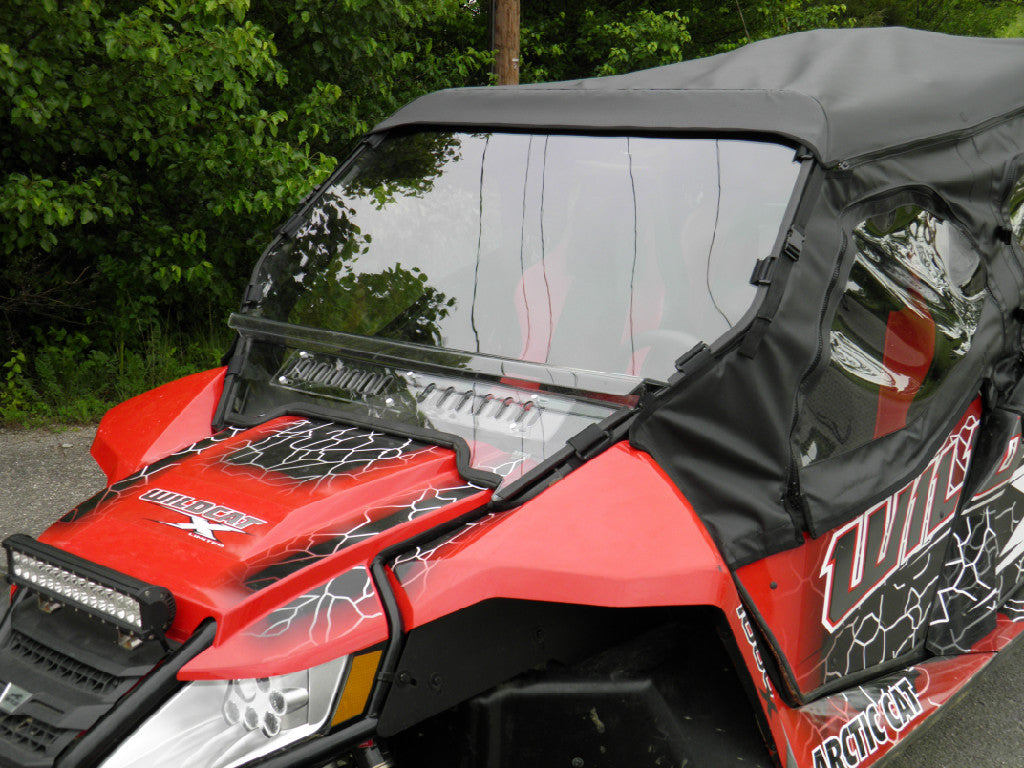 A close-up of a red and black Wildcat X featuring an Arctic Cat Wildcat Split Lexan Windshield (2012-2018) and partial soft top, parked on a paved surface with green trees in the background.