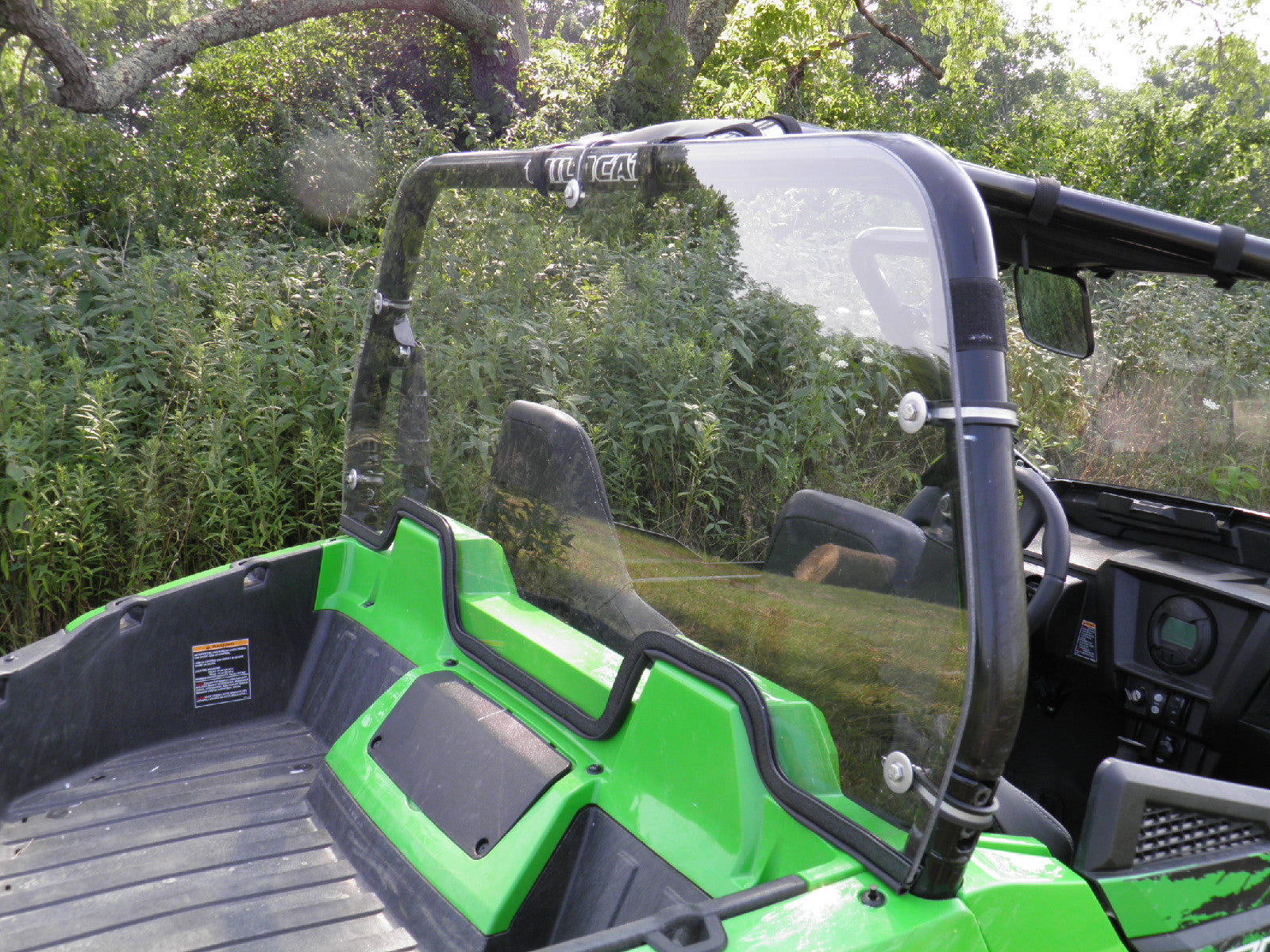 An Arctic Cat Wildcat Trail And Sport with a Lexan Back Panel is parked outdoors among trees and vegetation. Its interior, featuring black seats and dashboard, is visible through the windshield.