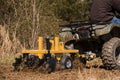 A person on an ATV pulls the Heavy Duty Commercial Grade Compact Disc through a field, tilling the soil. Churned earth marks the ground, mud covers the ATV wheels, and trees and tall grasses appear in the background.