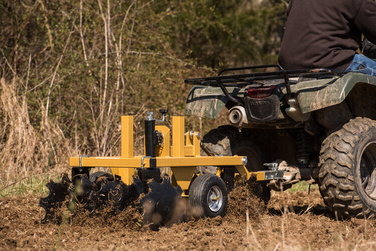 A person on an ATV pulls the Heavy Duty Commercial Grade Compact Disc through a field, tilling the soil. Churned earth marks the ground, mud covers the ATV wheels, and trees and tall grasses appear in the background.