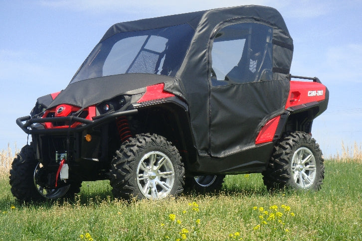 A Can-Am Commander (2014+) with a Full Cab Enclosure and Vinyl Windshield, in red and black, sits on green grass dotted with small yellow wildflowers. The off-road vehicle features large tires and clear side windows beneath a blue sky.