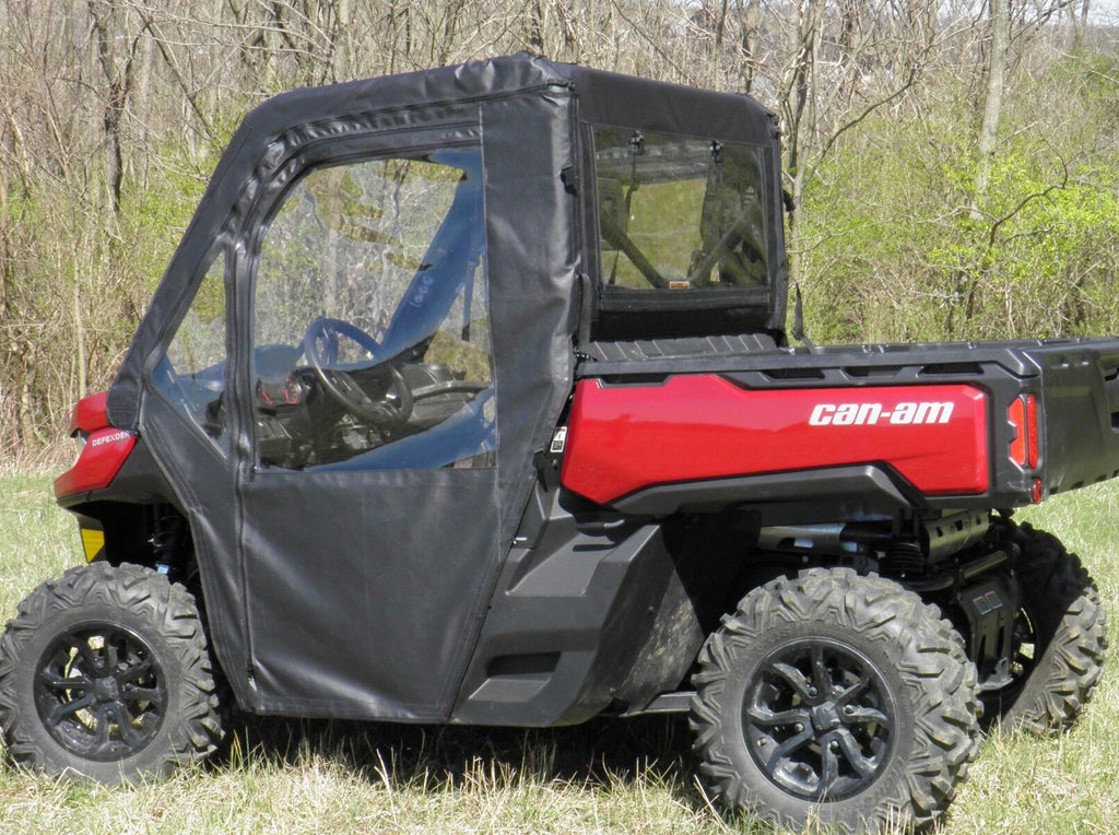 A red Can-Am side-by-side with rugged tires is parked on grass, featuring the Offroad Armor Can-Am Defender Soft Full Doors Kit with black soft doors and clear windows, set against sparse trees and green shrubbery.