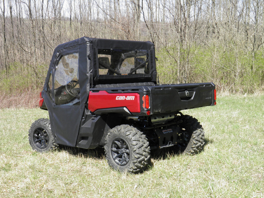 A Can-Am Defender equipped with Offroad Armor Soft Doors and Rear Window Combo is parked on grass near leafless trees, featuring a closed tailgate and large rugged rear tires for off-road use.