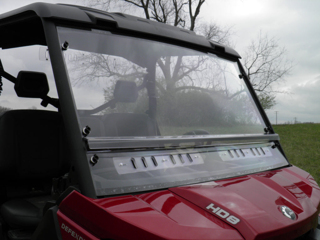 A close-up of the Offroad Armor Can-Am Defender Split Lexan Windshield installed on a red UTV parked on grass, showing its clear panels and secure mounting. Bare trees and a cloudy sky can be seen in the background.