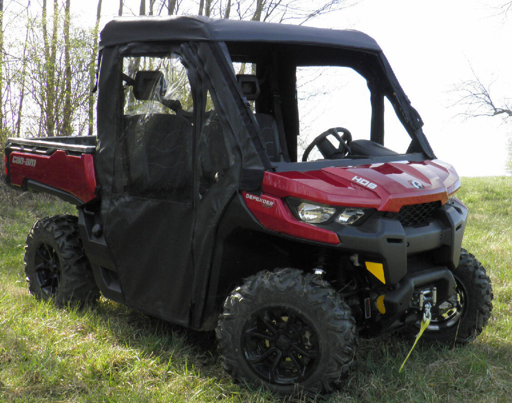 A Can-Am Defender with a Soft Full Enclosure for an existing hard windshield is parked on grass, featuring a black roof, partial side doors with clear windows, rugged tires, and a winch on the front bumper. Trees are visible in the background.