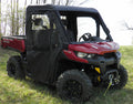 A Can-Am Defender with a Soft Full Enclosure for an existing hard windshield is parked on grass, featuring a black roof, partial side doors with clear windows, rugged tires, and a winch on the front bumper. Trees are visible in the background.