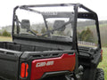 A Can-Am Defender with an Offroad Armor Lexan Rear Panel is parked on grass. The photo shows the rear view, highlighting the clear panel, two seats, roll cage, and red tailgate. Trees and rolling hills are in the background.
