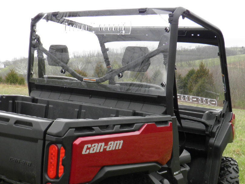 A Can-Am Defender with an Offroad Armor Lexan Rear Panel is parked on grass. The photo shows the rear view, highlighting the clear panel, two seats, roll cage, and red tailgate. Trees and rolling hills are in the background.