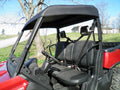 A close-up of a red utility side-by-side with an Offroad Armor Can-Am Defender Soft Top shows the open cabin—black seats, steering wheel, dashboard, and seatbelts under the black roof. Grass, trees, and a shed appear in the background.