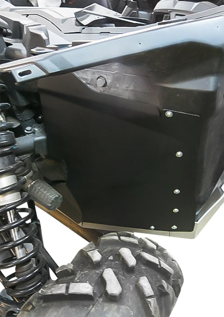 Close-up of a rugged off-road vehicle’s wheel and suspension. A black metal panel with bolts is mounted above the tire, partially enclosing the wheel well. The tire has a deep tread pattern and the visible suspension coil is shiny and robust.