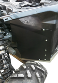 Close-up of a rugged off-road vehicle’s wheel and suspension. A black metal panel with bolts is mounted above the tire, partially enclosing the wheel well. The tire has a deep tread pattern and the visible suspension coil is shiny and robust.