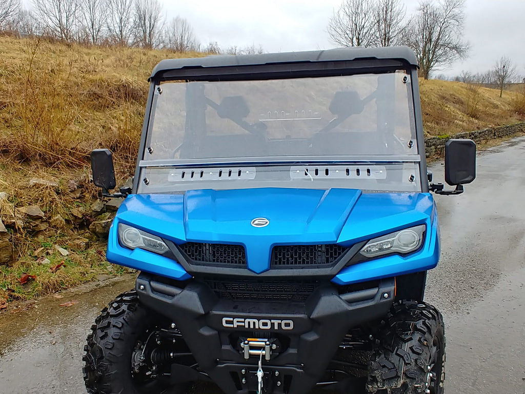 A Cf Moto Uforce 1000 with a Split Lexan Windshield (2019+) is parked on a wet road by dry grass and rocks. Its clear windshield, rugged tires, and front grill are visible, with bare trees and an overcast sky in the background.