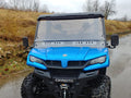 A Cf Moto Uforce 1000 with a Split Lexan Windshield (2019+) is parked on a wet road by dry grass and rocks. Its clear windshield, rugged tires, and front grill are visible, with bare trees and an overcast sky in the background.