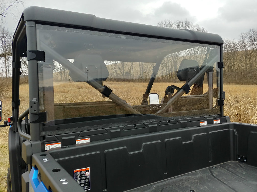 A rear view of a utility vehicle fitted with the Cfmoto Uforce 1000 Lexan Back Panel, featuring an empty black cargo bed, clear rear windshield, and headrests. Its parked in a grassy field with leafless trees under a cloudy sky; safety stickers visible.